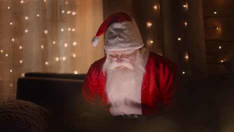 Porträt-Des-Weihnachtsmanns,-Der-Nachts-Im-Licht-Der-Weihnachtsbeleuchtung-Mit-Einem-Laptop-Arbeitet